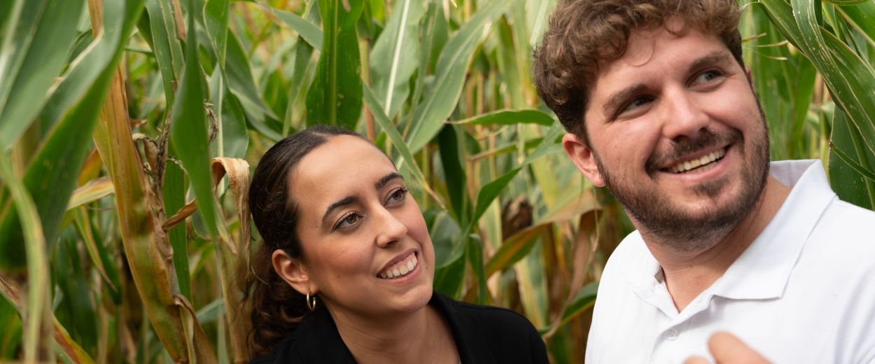 Cintia and Jesus F. engage in a field conversation among mature corn plants. They smile and look tow