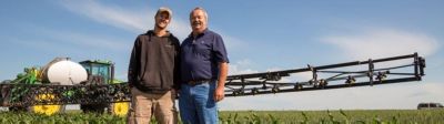 father and son in green crop field under blue sky with farm equipment in the background
