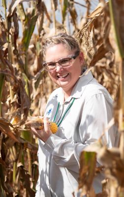 woman wearing Corteva white coat in cornfield holding an ear of corn