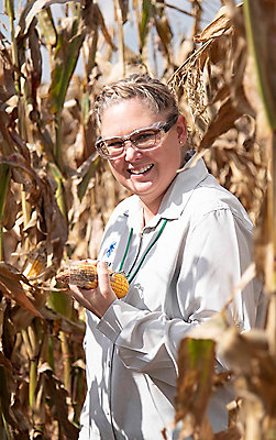 woman wearing Corteva white coat in cornfield holding an ear of corn