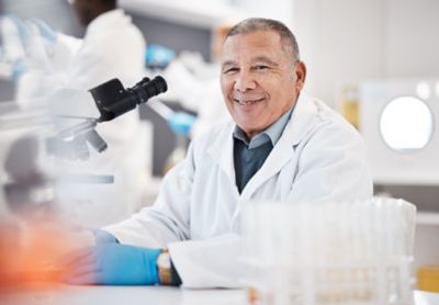 man in white lab coat in lab, seated at microscope
