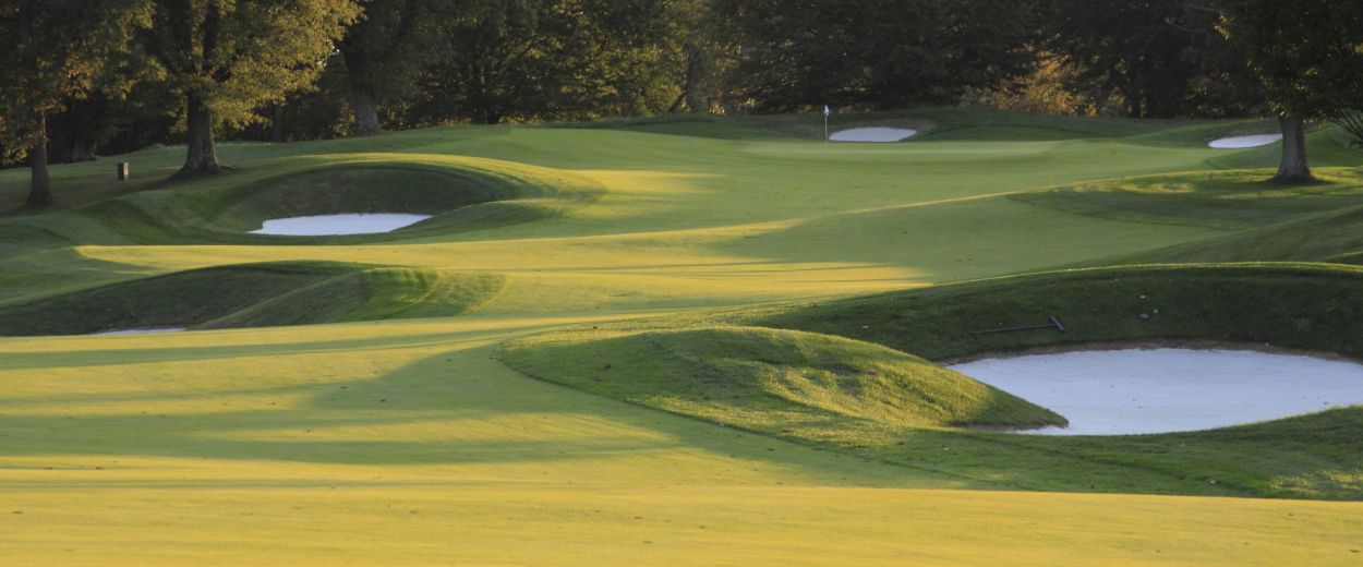 An empty golf course with rolling green fairways, several white sand bunkers and a distant flag, sur