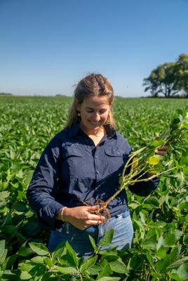 Valentina Luberriaga examines the roots of an uprooted soybean plant while standing in a green soybean field and wearing a Corteva Agriscience shirt. The image represents hands-on crop observation, plant health assessment and Corteva’s connection with agriculture.