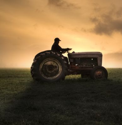 man driving tractor in field at sunset with dark yellow sky
