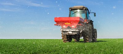 Trator agrícola aplicando adubação nitrogenada em lavoura verde sob céu azul, garantindo nutrição para híbridos de milho de alto desempenho.