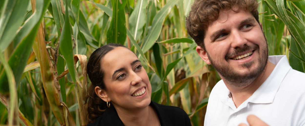 Cintia and Jesus F. engage in a field conversation among mature corn plants. They smile and look tow