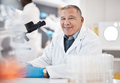 man in white lab coat in lab, seated at microscope