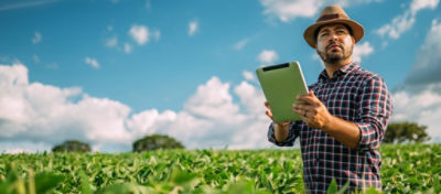 Agricultor usando um tablet para monitorar sua plantação em um campo verde, representando o uso de tecnologias digitais na agricultura moderna.