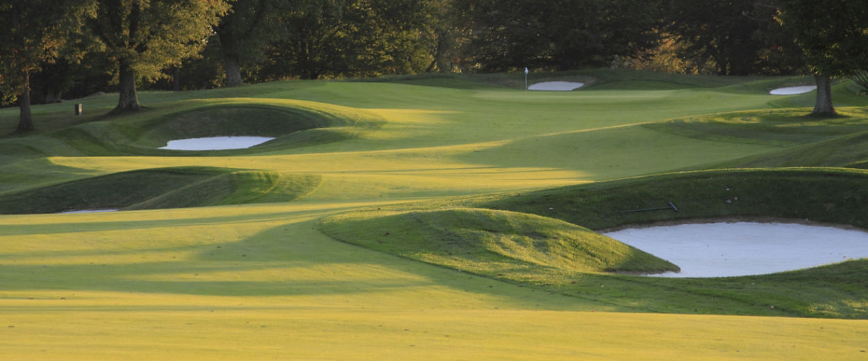 An empty golf course with rolling green fairways, several white sand bunkers and a distant flag, sur