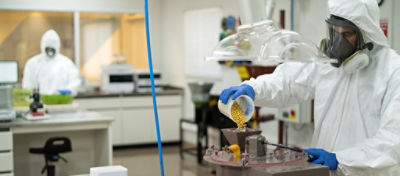 Profissional no laboratório introduzindo sementes de soja no equipamento. Ao fundo outro profissional trabalhando.