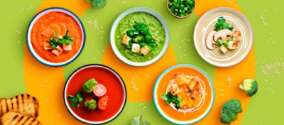 Five bowls of different types of soups with pieces of bread, broccoli and scallion in the background