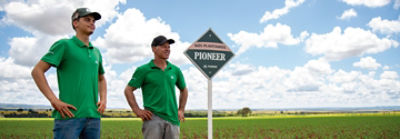 Pai e filho olhando para o horizonte com as mãos na cintura ao lado de uma placa escrito "Aqui Plantamos Pioneer".