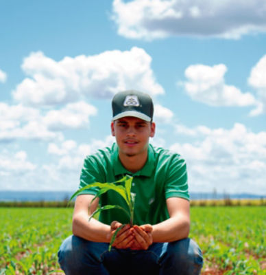 Homem agachado na lavoura de milho segurando uma planta na mão e olhando para a câmera.
