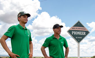 Pai e filho olhando para o horizonte com as mãos na cintura ao lado de uma placa escrito "Aqui Plantamos Pioneer".