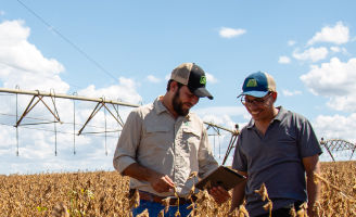 Dois homens de boné no meio de uma lavoura de soja olhando para a tela de um tablet.